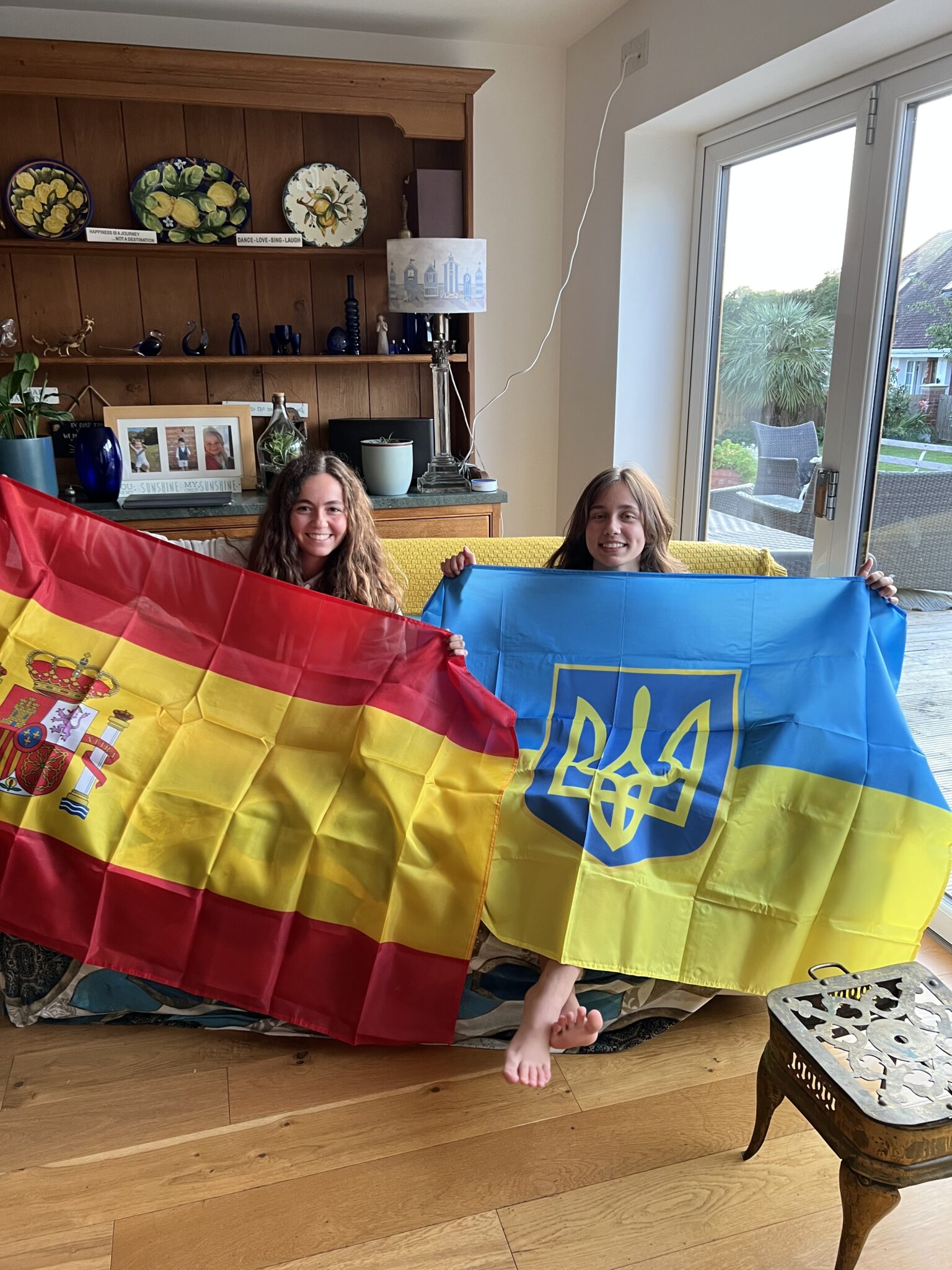 Two smiling teenagers standing indoors holding the flags of Ukraine and Spain.