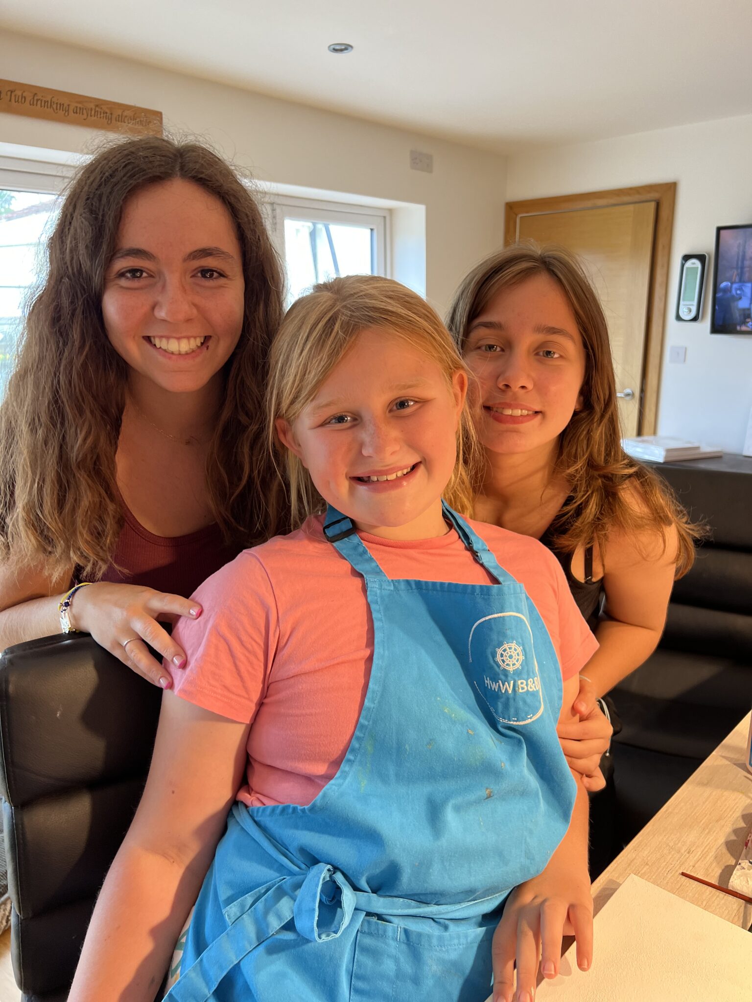 Three girls standing close together and smiling indoors; the youngest wears a blue apron.