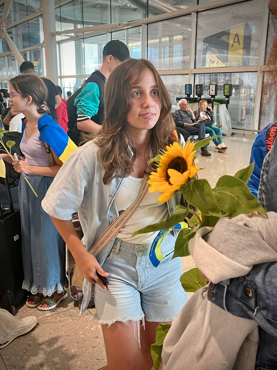 A teenage girl holding a bouquet of sunflowers inside an airport arrivals hall, surrounded by other passengers.